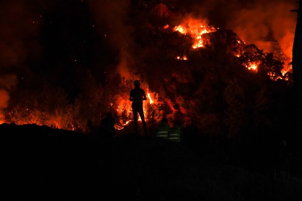 Dramática escena nocturna en la que tres siluetas de bomberos forestales observan las llamas devorar la vegetación del bosque. | Özgür Sürmeli, Pexels.